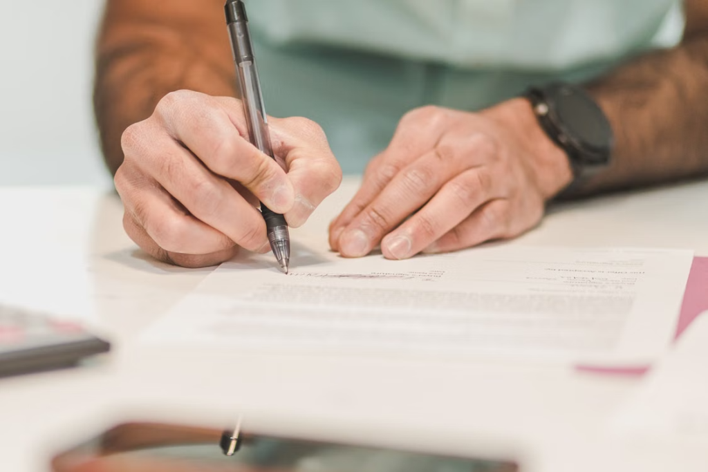 A person signs a form with a black pen while standing at a desk.