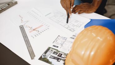A construction professional reviews blueprints and project charts with a hard hat on the desk.