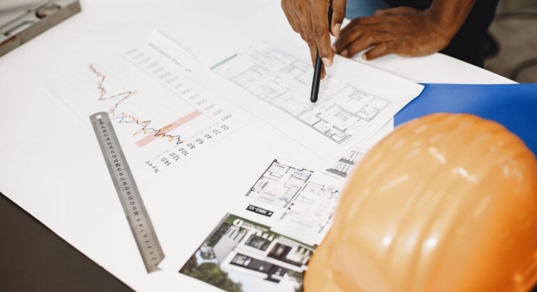 A construction professional reviews blueprints and project charts with a hard hat on the desk.