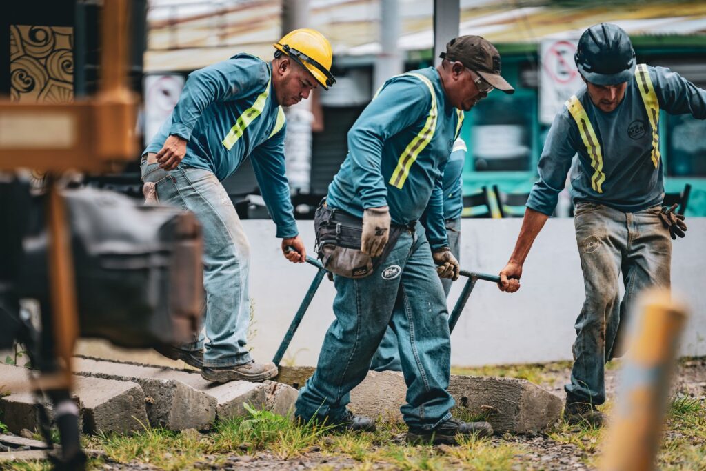 Three construction workers in matching high-visibility shirts and hard hats work together to carry a heavy metal frame across rough ground at a job site.
