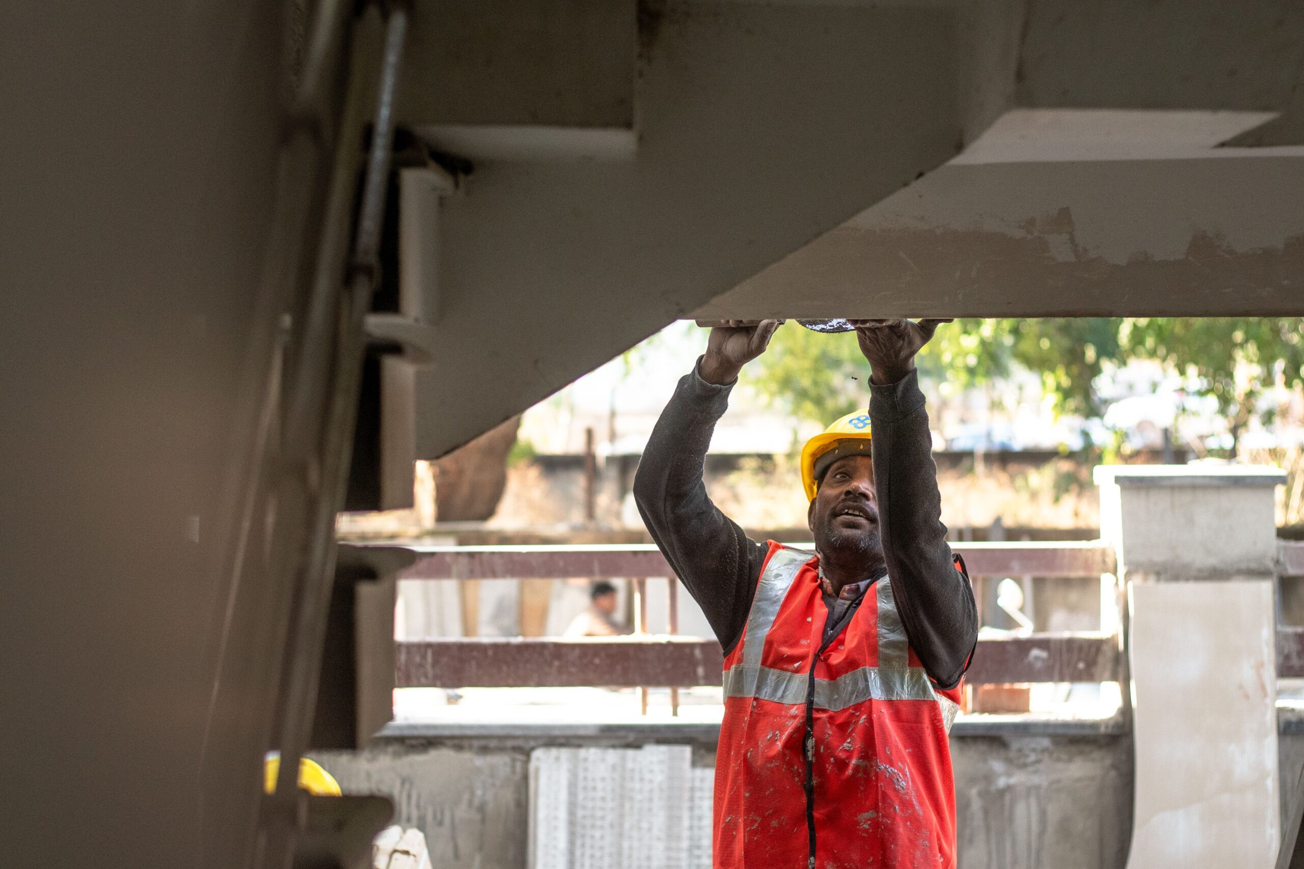 A construction worker in a hard hat and safety vest doing overhead work on a job site.